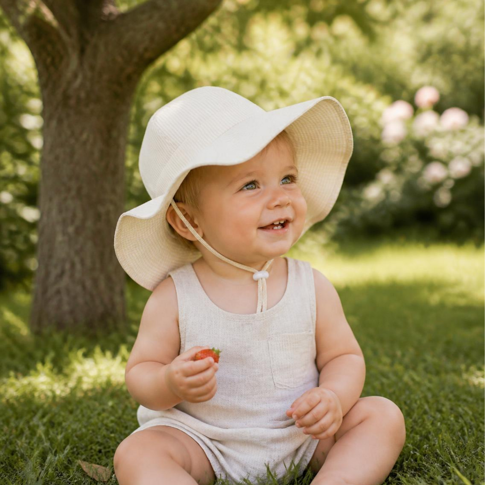Baby in a cream wide brim sun hat sitting in the shade of a tree